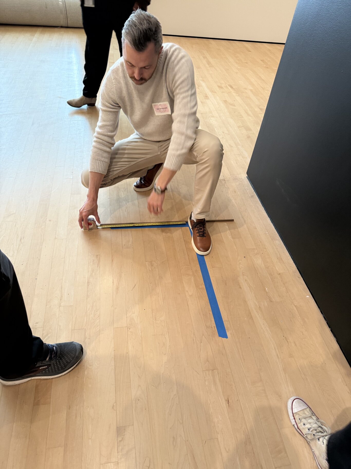Rob Itri Vincent, a Metí man in a light sweater, beige pants, and brown shoes is crouching while holding a tape measure stretched across the light wood floor. A blue tape line runs perpendicular to the measuring tape near a dark wall on the right. Other people's legs and shoes are partially visible around the scene.