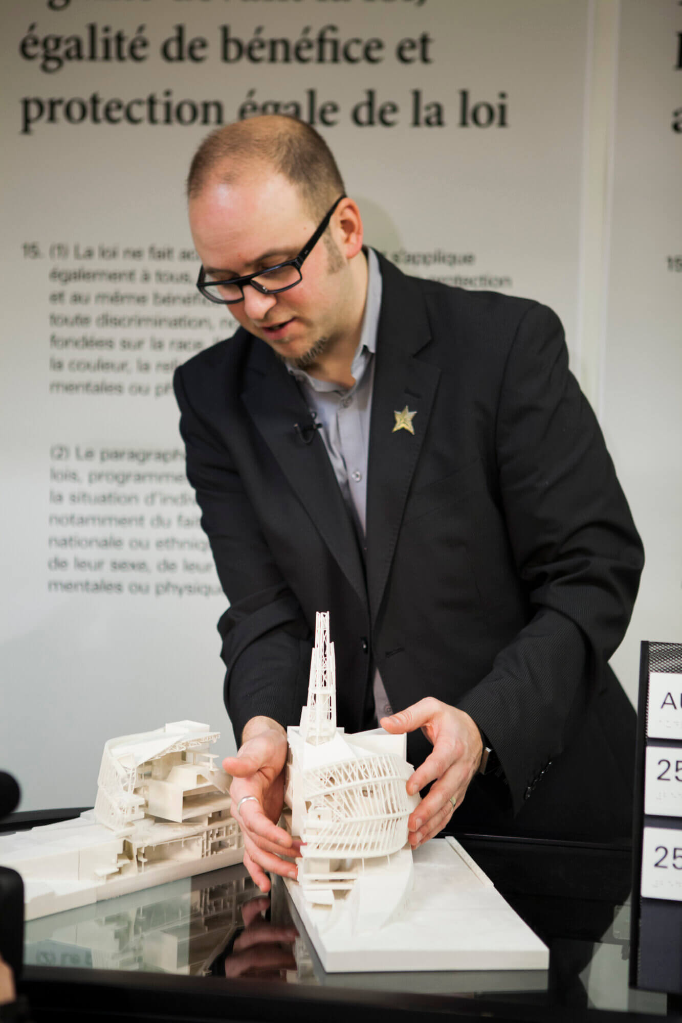 Corey Timpson, a white man wearing black-framed glasses, a dark blazer, leaning over a tabletop display, explaining a detailed white architectural scale model of a building. The model includes curved lattice-like structures and a tower-like element. His hands are positioned around the model as if demonstrating its features. Behind him is a large blurred wall or poster with French text, including phrases related to equality and legal protection.