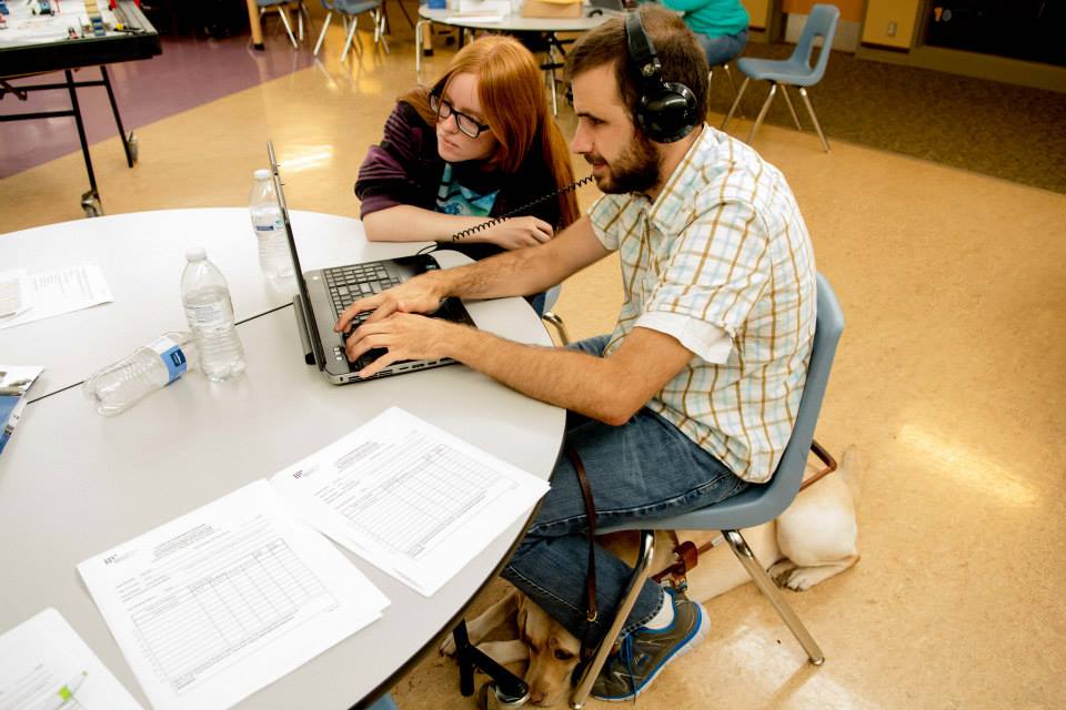 In a classroom, a man with light skin in a plaid shirt sits at a round table using a laptop and wearing large headphones. A red-haired woman with glasses leans close to watch the screen. The table has printed worksheets and water bottles scattered across it. A service dog, is lying or resting under the man's chair near his feet. Other chairs, tables, and people are visible in the background.