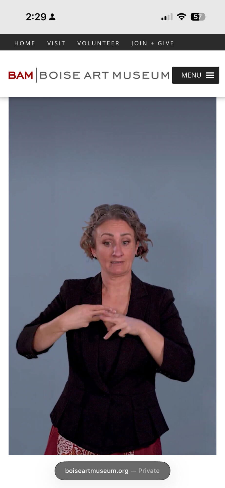 A smartphone screenshot of a video of a white woman signing on the Boise Art Museum website. At the top is a dark navigation bar with links reading "HOME," "VISIT," "VOLUNTEER," and "JOIN + GIVE." The museum branding appears underneath: "BAM | BOISE ART MUSEUM," with a black "MENU" button and hamburger icon on the right. The main content area shows a video frame of a white woman, Tara Adams, in a black blazer, positioned in front of a plain blue-gray background, mid sign. At the bottom, the browser bar displays "boiseartmuseum.org - Private."