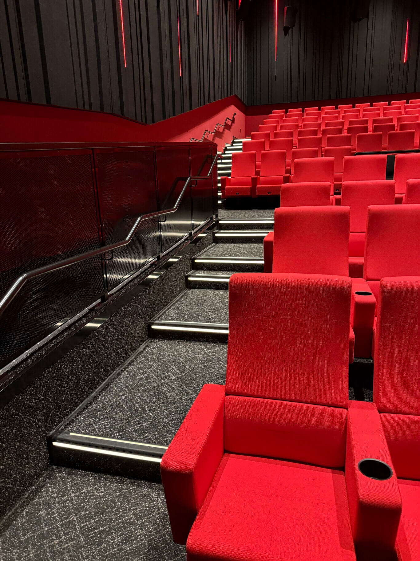 The interior of a movie theater or screening room. Rows of vivid red upholstered seats fill the right side and foreground, with cup holders built into the armrests. A stepped aisle runs through the seating area, featuring white illuminated strips along the stair edges for visibility. On the left is a black metal handrail and mesh barrier beside a walkway. The walls are dark gray or black with vertical acoustic paneling and narrow red light strips.