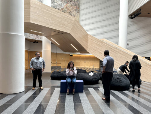 People sit and stand in a spacious contemporary building lobby and atrium. Several people are gathered in the lower area: Sina Bahram, a Persian man, stands near the center with his back to us and a long white cane in one hand, Maria Braswell, a white woman sits on a blue block looking at her phone connected into earphones, and others sit or stand near large black beanbag cushions. In the background, a large angular light-wood staircase and cuts diagonally across the scene, with built-in linear lights underneath. Tall white cylindrical columns frame the space, and the back wall has clean horizontal paneling. The floor features alternating gray and black striped tiles in a geometric pattern.