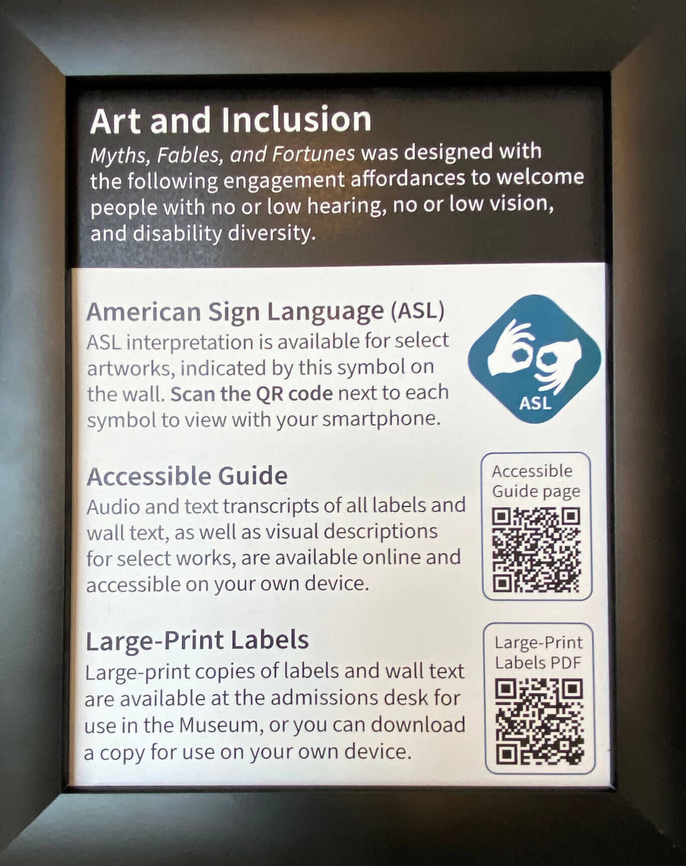 A close-up of a framed museum information sign. At the top, a dark header reads "Art and Inclusion." Below it, the sign explains that "'Myths, Fables, and Fortunes' was designed with the following engagement affordances to welcome people with no or low hearing, no or low vision, and disability diversity." The main white section lists accessibility resources. One section is titled "American Sign Language (ASL)" and says, "ASL interpretation is available for select artworks, indicated by a symbol on the wall. Scan the QR code next to each symbol to view with your smartphone." A blue ASL icon with hands making the sign for interpret appears to the right. The second section is titled "Accessible Guide" and says, "Audio and text transcripts of all labels and wall text, as well as visual descriptions for select works, are available online and accessible on your own device," with the image of a QR code beside it. The final section is titled "Large-Print Labels" and says, "Large-print copies of labels and wall text are available at the admissions desk for use in the Museum, or you can download a copy for use on your own device," with another QR code on the right.