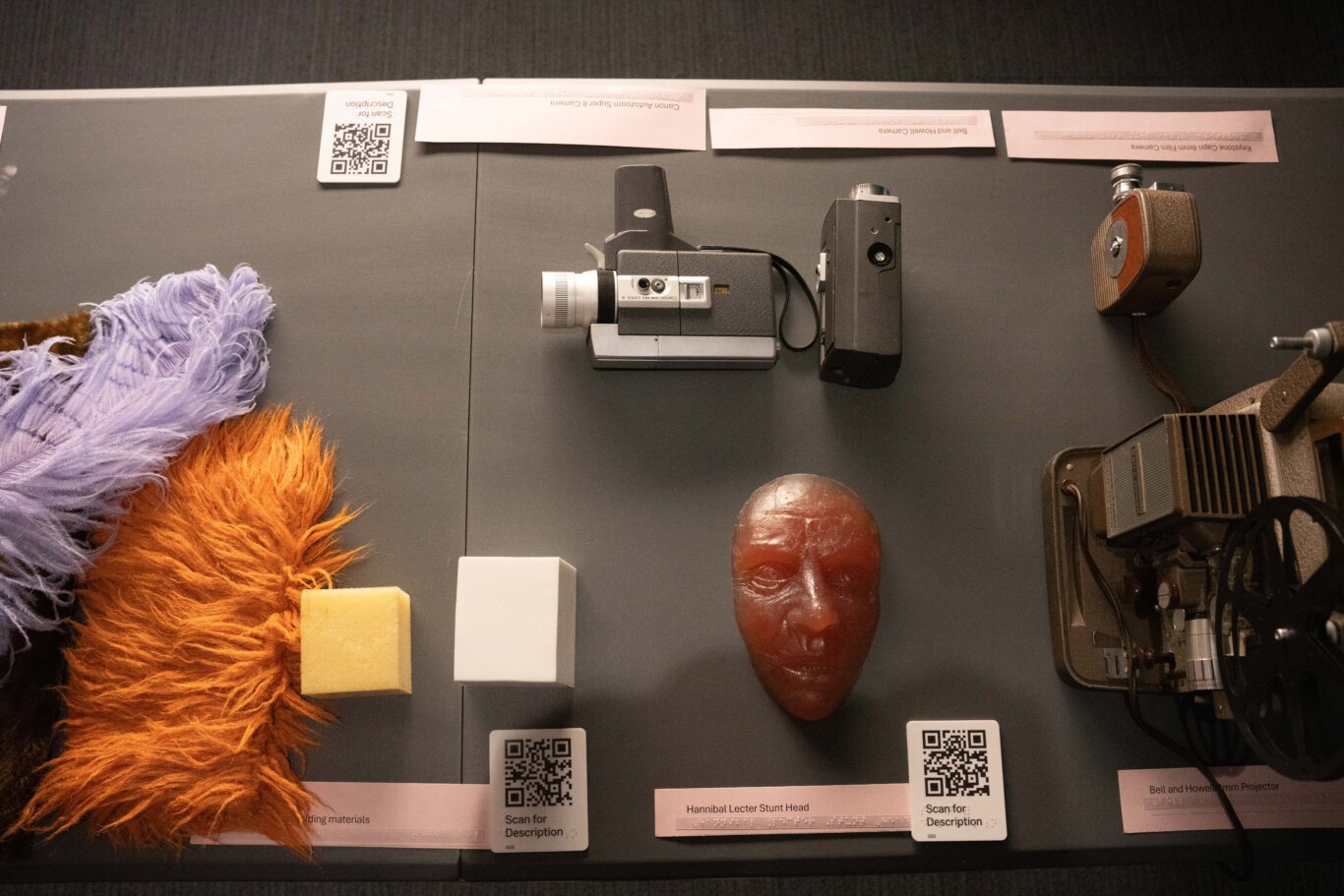 A table viewed from above, with several movie related objects with labels and QR codes arranged on a dark surface. The items are laid out along the top and bottom of the table with labels underneath in both print and Braille next to a tactile QR code that says Scan for Description in printed text and Braille. Starting at the bottom edge of the table, on our left, are colorful muppet crafting materials, including shaggy orange fur, pale purple feathers, and foam blocks. In the midle is a reddish-brown molded head labeled "Hannibal Lecter Stunt Head." On our right is a large vintage Bell and Howard Film Projector. On the other side of the table are a row of vintage film devices, including a Canon Autozoom Super 8 camera.