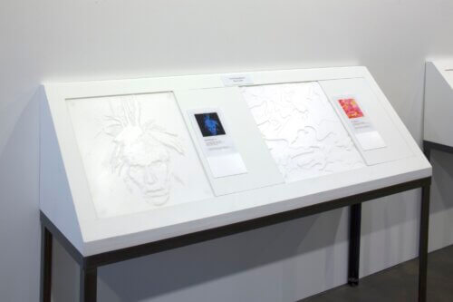 A slanted white museum or gallery display table against a plain wall. The display is labeled "Tactile Reproductions Please Touch," suggesting it is designed for hands-on accessibility. On the left is a large pale raised-relief image resembling a portrait, while the center includes a label card with a small blue-toned self-portrait image. On the right are additional raised outline patterns and a colorful printed artwork label. The display sits on black metal legs in a clean, minimal exhibition space.
