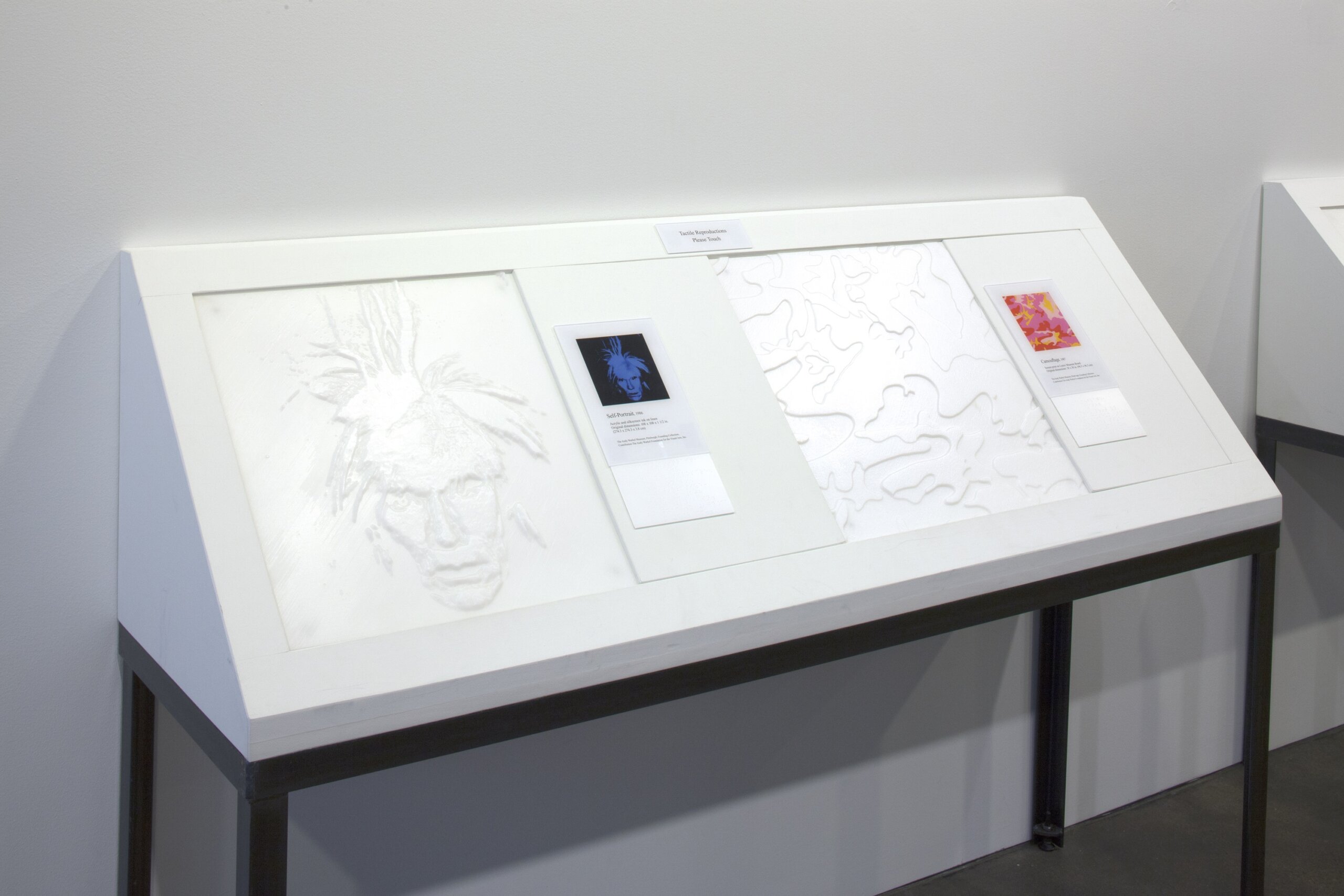 A slanted white museum or gallery display table against a plain wall. The display is labeled "Tactile Reproductions Please Touch," suggesting it is designed for hands-on accessibility. On the left is a large pale raised-relief image resembling a portrait, while the center includes a label card with a small blue-toned self-portrait image. On the right are additional raised outline patterns and a colorful printed artwork label. The display sits on black metal legs in a clean, minimal exhibition space.