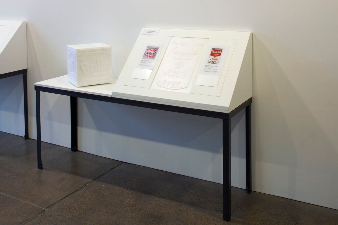 A gallery or museum installation featuring tactile reproductions of iconic pop-art consumer packaging. On the left table is a white embossed cube resembling a Brillo soap pads box. On the larger angled display to the right are mounted panels with raised white outlines and small printed reference images, including a Campbell's Cream of Mushroom soup can and a Brillo box. A small sign at the top reads "Tactile Reproductions Please Touch." The display sits against a light-colored wall on white tables with black metal legs, with a concrete floor beneath.
