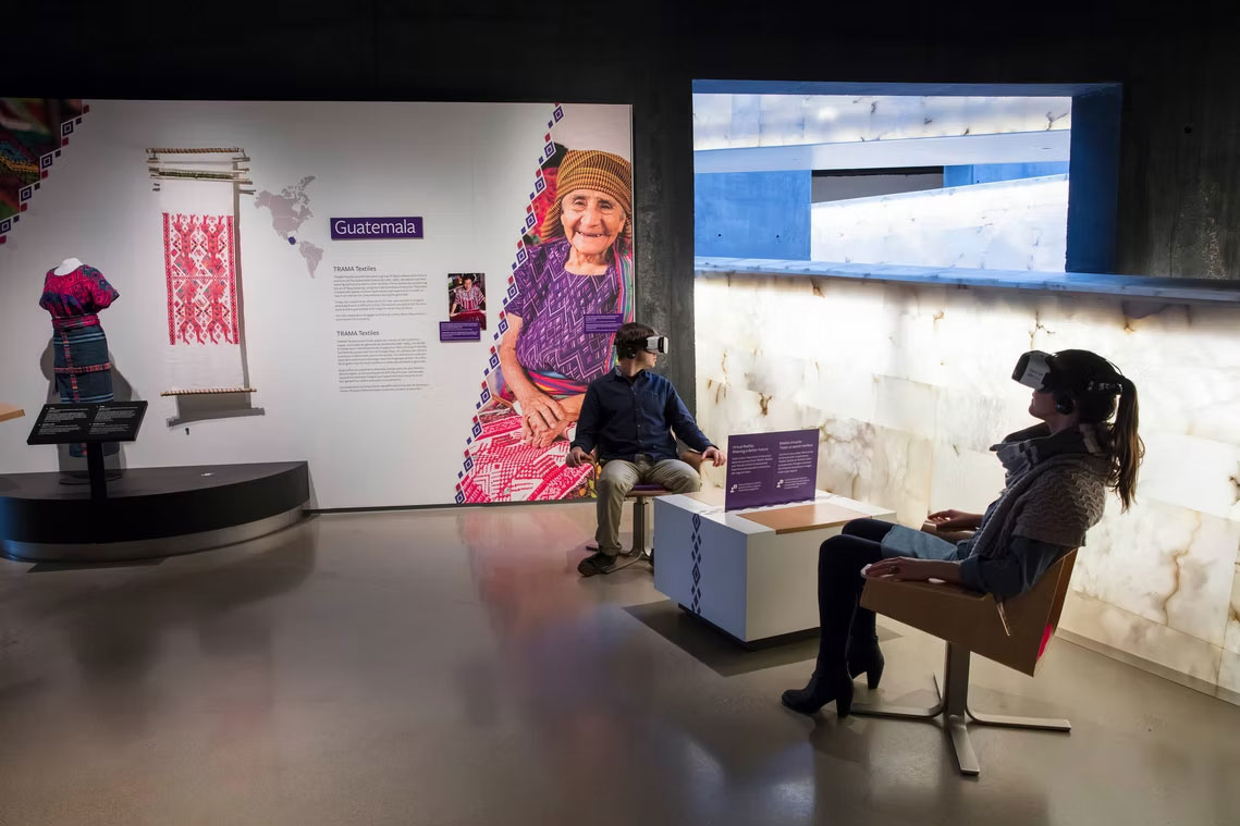 A museum exhibit featuring content about Guatemalan weaving practices. On our left, there is a display about Guatemala with traditional woven textiles, a mannequin dressed in colorful clothing, a loom with a weaving, informational panels, and a large photograph of a smiling older woman with wrinkled light brown skin tone in traditional Guatemalan attire. On our right, two visitors sit in chairs wearing virtual reality headsets, appearing to experience an interactive exhibit. The room is dimly lit with illuminated walls and display panels.