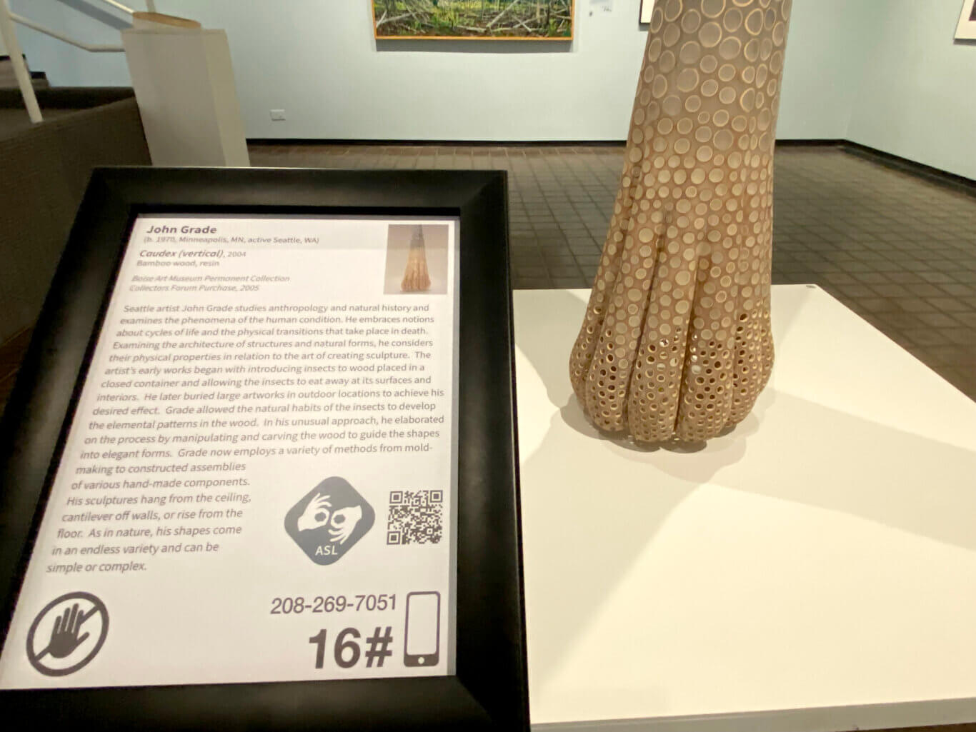 A gallery setting shows a black-framed exhibit label with text identifying the artist as John Grade and the work, partially visible in the background, as "Caudex (vertical)," 2004, made from bamboo wood and resin. The label includes a description of the artist's process, a small image of the sculpture, an ASL icon, a QR code, and an audio/phone guide number "208-269-7051" with the number "16." To the right, part of the actual sculpture is visible: a tall, vertical, beige form covered with many circular holes or ring-like perforations. In the background are gallery walls, a staircase or ramp area, tiled flooring, and framed artwork on the wall.
