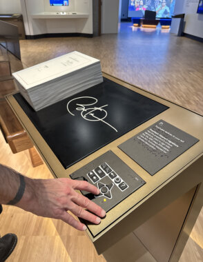An interactive museum exhibit with a stack of paper's and large tactile white signature of Barack Obama. On our left of the objects is a person's hand with light skin tone using a UXP, a tactile control panel, with raised buttons, directional arrows, volume controls, zoom buttons, and a help button all labeled in Braille. Above the UXP are is a label in print and Braille reading, "Touchable replicas to your left. President Obama's signature. Feel the presidents unique signature, used to sign bills like the 906 -page Affordable Care Act: Activate keypad for a guided description." raille labels and printed instructions describing touchable replicas related to President Obama's signature.