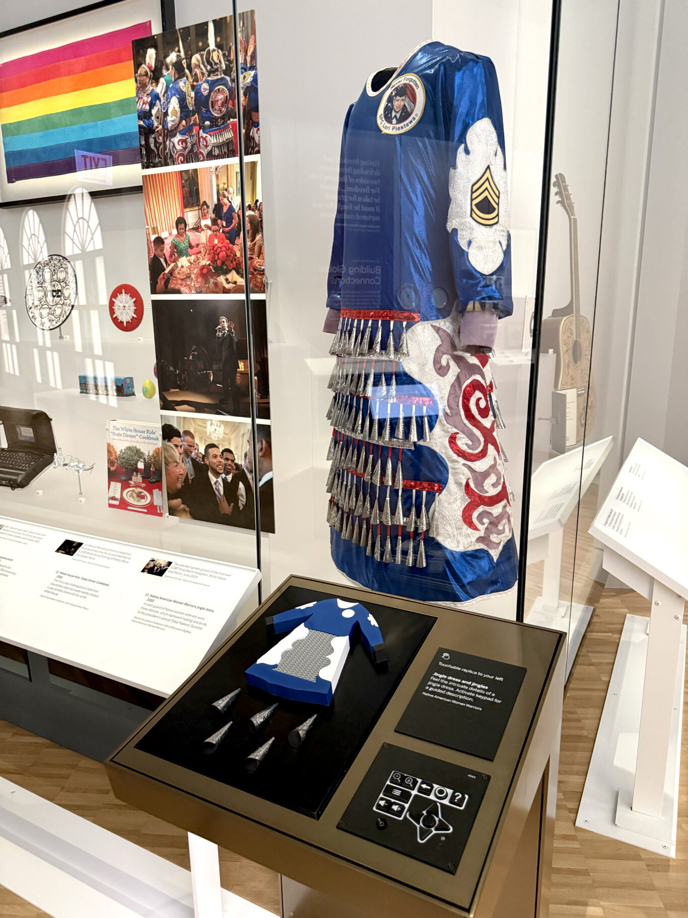 A well-lit museum gallery with multiple artifacts and photographs arranged inside glass display cases. On the right, a striking blue satin-like jingle dress is displayed on a mannequin, decorated with silver metal cones, red trim, white embroidered panels, and a patch honoring "SPC Lori Piestewa." In front of it is an interactive station with a tactile replica of part of the dress, loose cone-shaped jingles, and the UXP, a tactile keypad, with accessibility controls for guided description. On the left side of the exhibit are a rainbow pride flag, photos of people at events including dancers and White House gatherings, and other objects such as a decorative plate, a small red circular item, a laptop, and a cookbook.