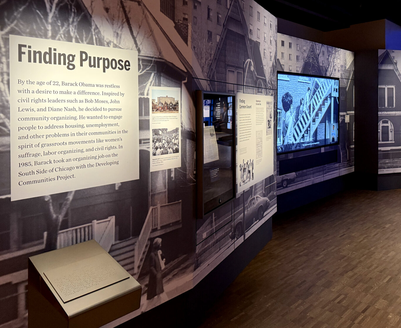 A dimly lit museum gallery with a large black-and-white photographic mural of vintage urban buildings covering the walls. A prominent exhibit panel reads "Finding Purpose" and describes Barack Obama, at age 22, becoming interested in community organizing and addressing issues such as housing and unemployment on Chicago's South Side. In the foreground, a low pedestal holds a tactile Braille panel with the same text. Additional panels include historical photographs, text about organizing on the South Side, and displays related to civil rights and grassroots movements. Several embedded screens show video or multimedia content, including a blue-toned video on the right.