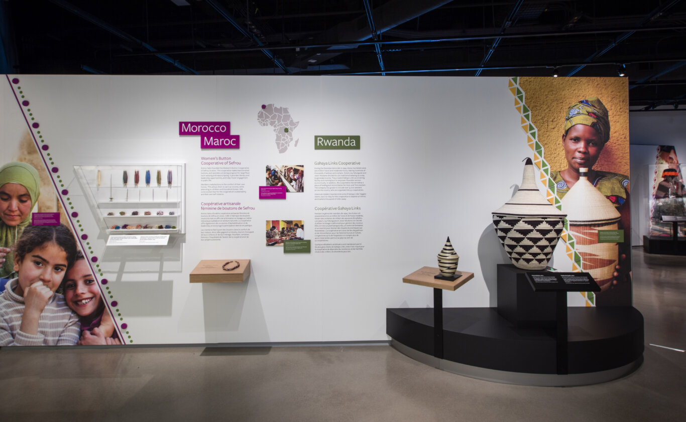 A museum exhibit focused on Morrocan and Rawandan artisan cooperatives. Large images of artists, baskets, jewlery, wall text, and wall graphics label sections for "Morocco" and "Rwanda," with a pale map of Africa marking locations. The Morocco section on our right discusses the Women's Button Cooperative of Sefrou and includes a glass case with colorful handmade buttons or textile-related objects, a ledge with a necklace that can be touched, along with large portraits of women and children with medium light skin tone smiling at us. The Rwanda section on our right highlights the Gahaya Links Cooperative, small photos of artisans working, and two woven baskets similar in shape and design but different in size displayed on pedestals. behind the basket is a woman with medium dark brown skin tone wearing a head wrap that matches her shirt. The exhibit uses white walls, magenta and green labels, geometric decorative borders, and large portrait images, while the dark exposed ceiling and spotlights.
