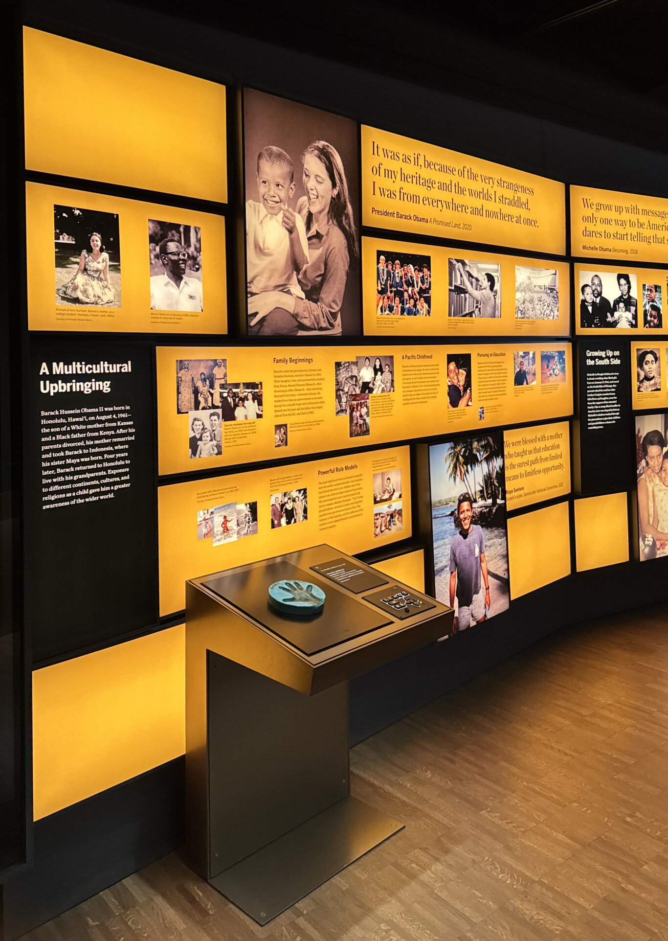 A dimly lit museum gallery featuring a large wall display in black and vivid yellow labeled "A Multicultural Upbringing." The exhibit includes historical photographs, biographical text panels, and quotes related to Barack Obama and Michelle Obama. A prominent section titled "A Multicultural Upbringing" describes Obama's birth in Honolulu and his family background, while other panels discuss "Family Beginnings," "A Pacific Childhood," and education. Large photos show Obama as a child with family members, alongside smaller archival images. In front of the wall is an interactive display table of a small tactile handprint and a UXP, a tactile keypad.