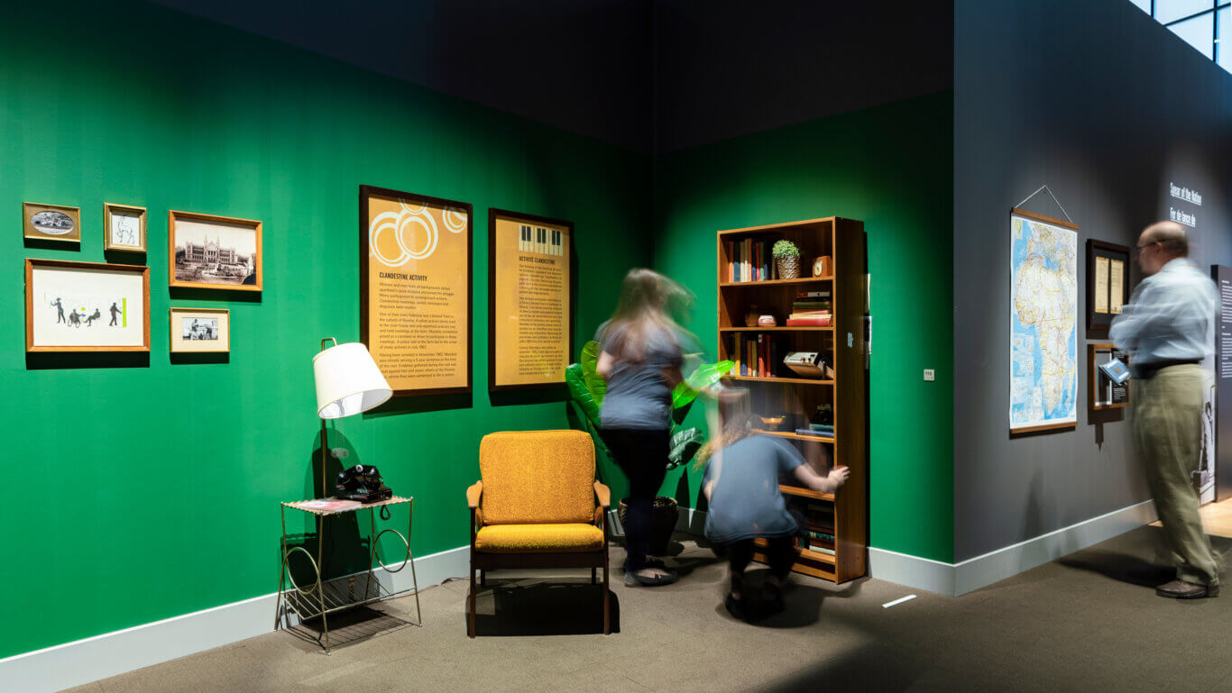 An indoor museum or exhibition gallery of an old office like space with vivid green walls and dark gray accent sections. Several framed photographs and documents hang on the left wall, while large informational panels in the center discuss "Clandestine Activity," including text in English and French. The exhibit is styled like a historical room, featuring a mustard-yellow armchair, a small side table with a white lamp and black rotary telephone, a tall wooden bookshelf filled with books and objects, and a wall-mounted map. A few visitors appear blurred from motion, suggesting they are walking through or interacting with the display. Bright overhead lighting creates dramatic shadows and highlights across the walls and objects.