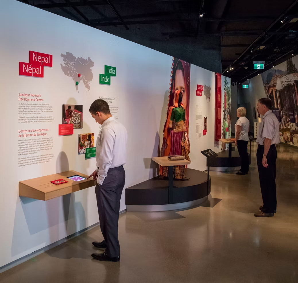Three people with light skin tone wearing business casual clothes stand in an exhibition with large wall panels focused on Nepal and India. On the wall from left to right are red and green labels that read "Nepal - Janakpur Women's Development Center" and "India - Self Employed Women's..." with the rest of the text cut off behind a visitors head. The exhbiti contains a map, text sections, photos, and a digital touch screen with fabric touch sample. Farther down the gallery on our right, two other visitors are looking at additional exhibits of colorful clothing and fabrics. The space has polished floors, dark ceilings with track lighting, and a modern museum layout.