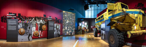 A dramatic indoor museum display focused on mass mobilization and anti-apartheid struggle. On our left, large red walls are covered with archival photos and black-and-white silhouettes of crowds of Black South African protesters holding rusty trash can lids, with prominent bilingual text such as "Mass Mobilization" and "What Freedom?" Across the room stands a massive yellow armored tank, towering twice as high as the protestors. Around it are tall dark information panels with historical text, photographs, posters, and archival imagery. Through the middle of the room on the floor is a white strip of raised tactile floor markers.