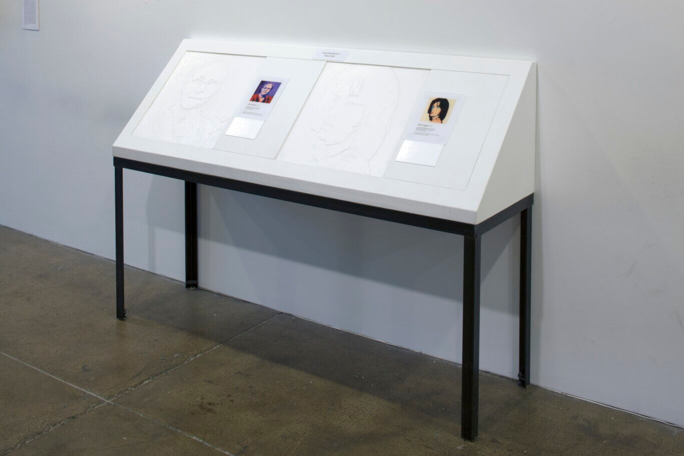 An art gallery installation: a large white, angled display case on black metal legs stands against a plain white wall on a polished concrete floor. Inside the case are two very light, raised or embossed portrait drawings, designed like tactile reproductions. Each portrait is paired with a small printed color image and explanatory label; the visible labels appear to reference Julia Warhola and Mick Jagger. A small sign at the top of the case reads "Tactile Reproductions Please Touch," indicating that visitors are invited to feel the artworks.