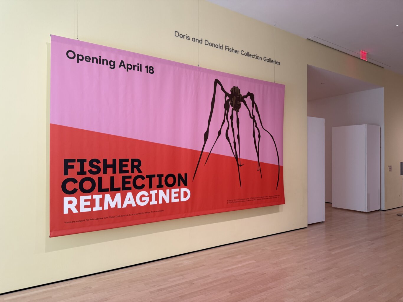 An interior museum spacewith a large suspended exhibition banner dominating the scene. The banner is pink on top and red on the bottom with bold black and white text on the banner which reads "Opening April 18" and "Fisher Collection Reimagined." On the right side of the banner is a black silhouette illustration resembling a spider sculpture. Above the banner, wall text identifies the space as the "Doris and Donald Fisher Collection Galleries." A red exit sign is visible in the background near the ceiling.