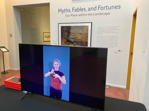 An indoor museum or gallery space with someone signing on a large TV in front of the entrance to an exhibit. To the right of the entrance on the wall, large blue text reads "Myths, Fables, and Fortunes," with the subtitle "Our Place within the Landscape" beneath it. Below and beside the title are exhibit text panels and a framed landscape artwork showing a river and trees. In the foreground, a large flat-screen TV sits on a table, displaying a white woman with pale skin tone, set against a blue background signing the word, "interpret." On the table are a small stack of red-framed tablets with handles. In the background, on our left, an opening leads into another gallery area with a small framed sign describing access information.
