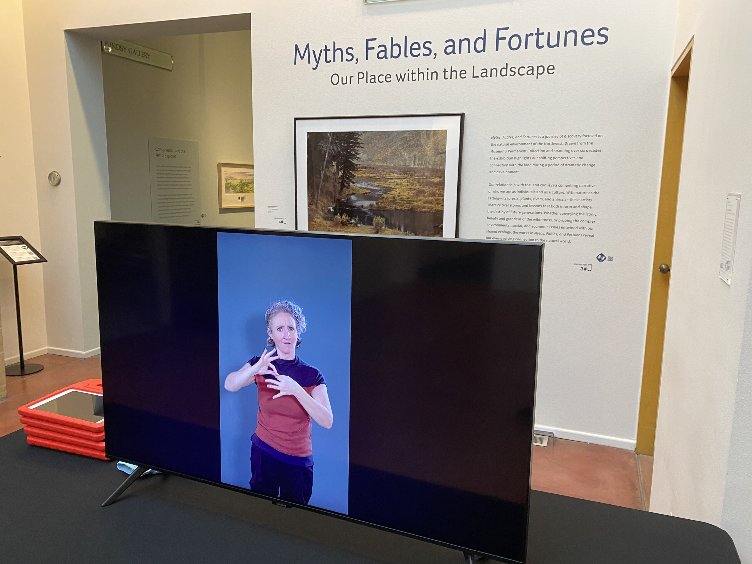 An indoor museum or gallery space with someone signing on a large TV in front of the entrance to an exhibit. To the right of the entrance on the wall, large blue text reads "Myths, Fables, and Fortunes," with the subtitle "Our Place within the Landscape" beneath it. Below and beside the title are exhibit text panels and a framed landscape artwork showing a river and trees. In the foreground, a large flat-screen TV sits on a table, displaying a white woman with pale skin tone, set against a blue background signing the word, "interpret." On the table are a small stack of red-framed tablets with handles. In the background, on our left, an opening leads into another gallery area with a small framed sign describing access information.