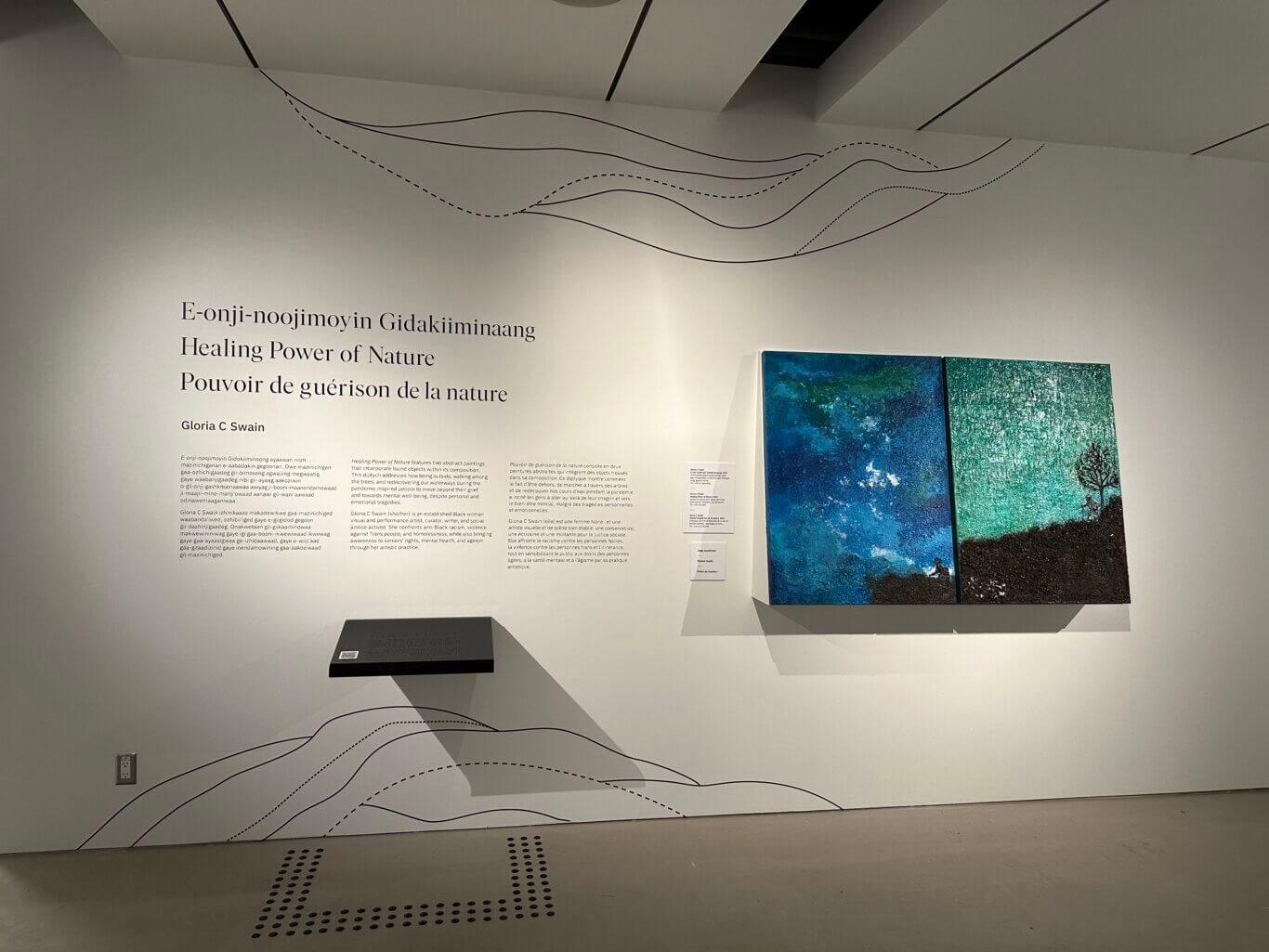 An art gallery installation on a white wall. Large serif text introduces the exhibition, "Healing Power of Nature," in three languages, Anishinaabemowin, English, and French, followed by the artist's name, Gloria C Swain. Beneath the title are blocks of explanatory wall text in multiple languages. Underneath the text is a black reader rail protruding from the wall, with tactile floor markers below it on the gray floor. On our right are two abstract highly textured paintings, one predominatly blue and the other predominatly green. Black solid and dashed wavy lines run across the wall and ceiling like flowing water or pathways.