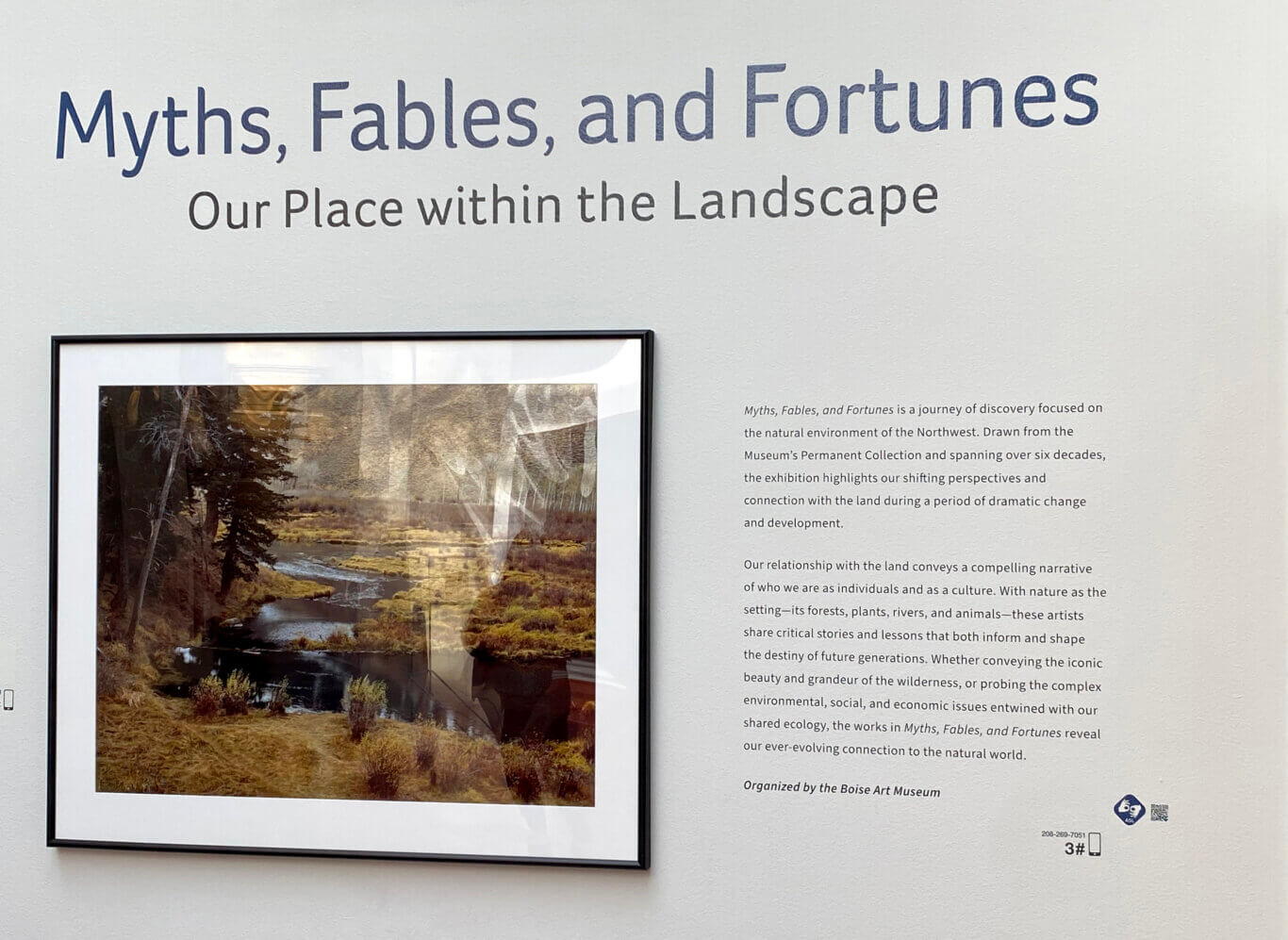 A white museum wall with large blue-gray lettering reading "Myths, Fables, and Fortunes," with the subtitle "Our Place within the Landscape" beneath it. Below the title is a framed landscape image featuring trees, wetlands, water, and earthy vegetation tones. On our right, a block of exhibition text explains that the show explores the natural environment of the Northwest, changing perspectives on land, and humanity's connection to nature. The text notes that the exhibition is organized by the Boise Art Museum. In the lower corner on our right are a cluster of icons: the symbol for ASL interpration, a QR code, a smart phone, a phone number, and the number three.