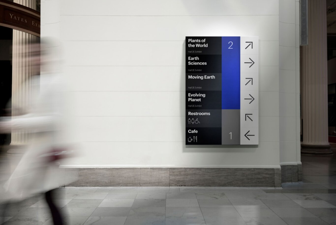 An interior museum or gallery corridor with a large modern wayfinding sign mounted on a pale wall. The sign lists the destination on the our left, "Plants of the World," "Earth Sciences," "Moving Earth," "Evolving Planet," "Restrooms," and "Cafe," with arrows indicating directions and floor numbers 1 and 2 on our right. The destinations are written in clear white text on a black background with ifons below the restrooms and cafe. To the right is a blue colum with the number two for the exhibits and a grey column underneath it with the number one for the restrooms and cafe. On the your right is the final column in the sign with large black arrows on a white background. The space has stone flooring, tall columns, and a clean institutional design. A person in a light-colored coat is motion-blurred while walking across the left side of the frame.