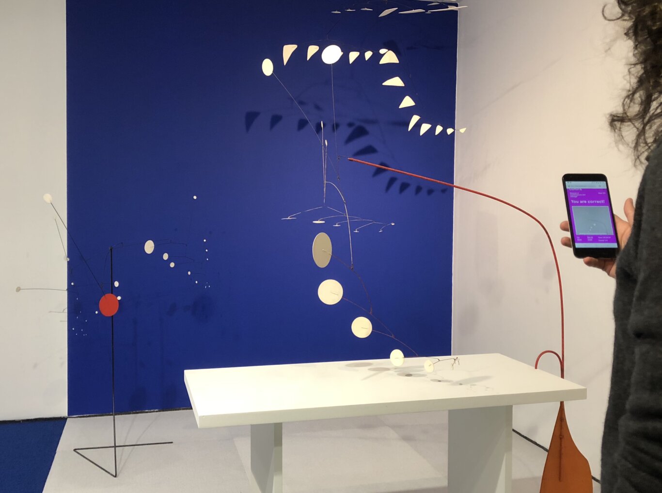 A large deep blue wall forms the backdrop for several abstract kinetic sculptures or mobiles made of thin wires and small suspended shapes in an exhibition. Many of the pieces are cream or white, with one bright red circular disk near the lower left and an orange cone-like object at the far right. A white display table sits in the foreground with more small mobile elements casting shadows. On the right edge, part of a person with curly hair and light skin tone is visible holding a smartphone; the phone screen shows a purple interface with the message "You are correct!" A red cable or rod crosses diagonally through the scene.