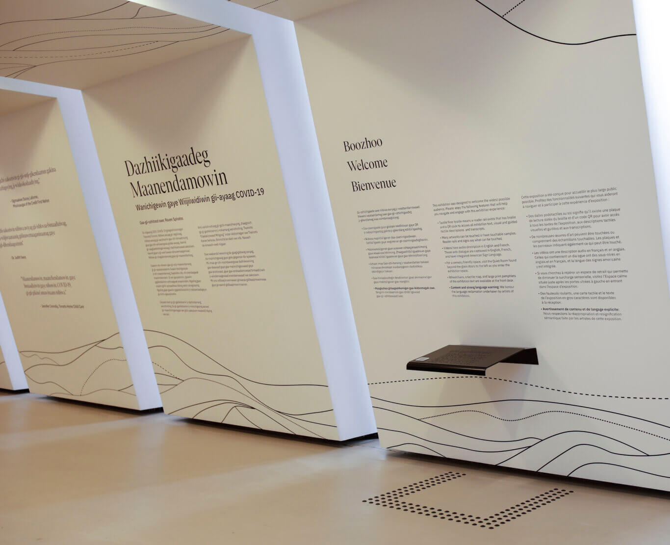 A modern exhibition space with white wall panels separated by narrow deeply recessed windows. Large printed text appears on the walls in multiple languages, including Anishinaabemowin, English, and French. On the right panel, the words "Boozhoo," "Welcome," and "Bienvenue" are prominently displayed above paragraphs explaining accessibility features for the exhibition, such as tactile floor braille, QR codes, audio description, captions, tactile samples, wheelchairs, and large-print pamphlets. On our right wall, a reader rail with Braille and a QR code sits with rows of dark raised tactile dots framing it's edge on the floor below. The panels are decorated at the bottom with thin black wavy and dotted lines, resembling map contours or pathways.