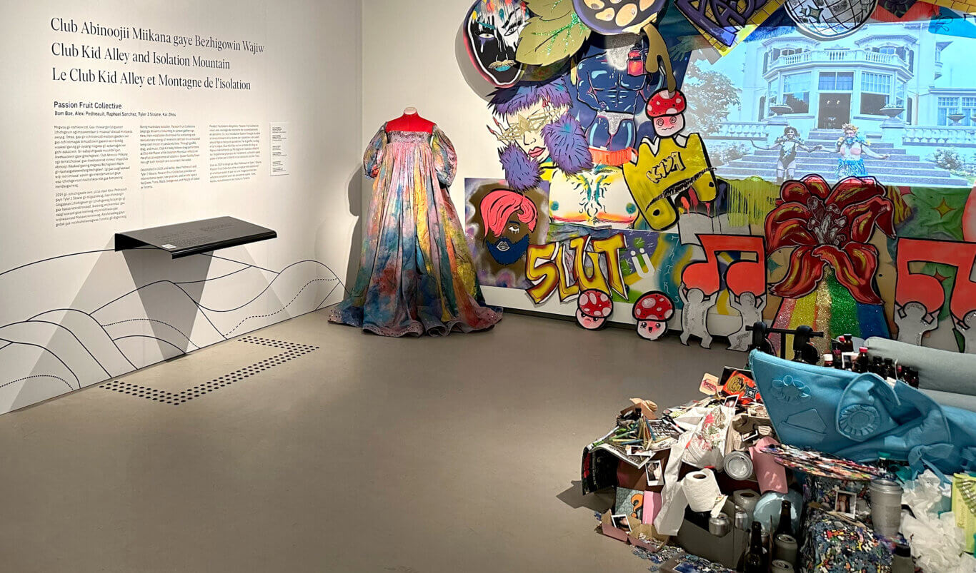 A contemporary art exhibition space with white gallery walls and a large installation. On the wall on our left, large text introduces the work in three languages, Anishinaabemowin, English and French. The heading says, "Club Kid Alley and Isolation Mountain," with the artist group listed as Passion Fruit Collective. Beneath the title are blocks of explanatory wall text and a black reader rail with Braille and a QR code framed on the grey floor by black tactile floor markers. In the center stands a mannequin wearing a dramatic, floor-length, multicolored dress with voluminous sleeves. To our right, the installation becomes a dense installation: bright painted grafiti cutouts of cartoon-like faces, mushrooms, bold lettering, glittery elements, and costume-like shapes overlap in front of a projected image of a large building and performers. In front of the screen in the foreground is an installation of a blue couch consumed by a cluttered assemblage of props, puzzle pieces, bottles, Covid-19 supplies, and polaroids.