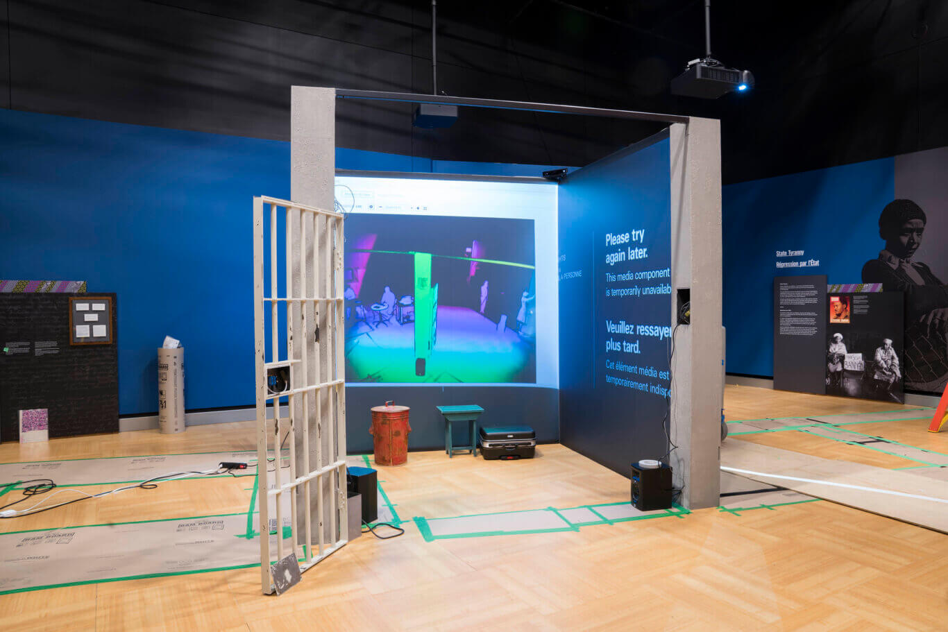 An indoor exhibition space with an installation under construction on a wood floor. In the center is a partial concrete-and-metal structure resembling a prison cell, with a white barred gate standing open. A projector mounted overhead casts a colorful video or digital scene onto a wall inside the structure. Nearby are speakers, cables, a suitcase, a small stool, and a red barrel. The surrounding walls are painted bright blue and dark gray, with large text including "Please try again later. This media component is temporarily unavailable" in English and French.