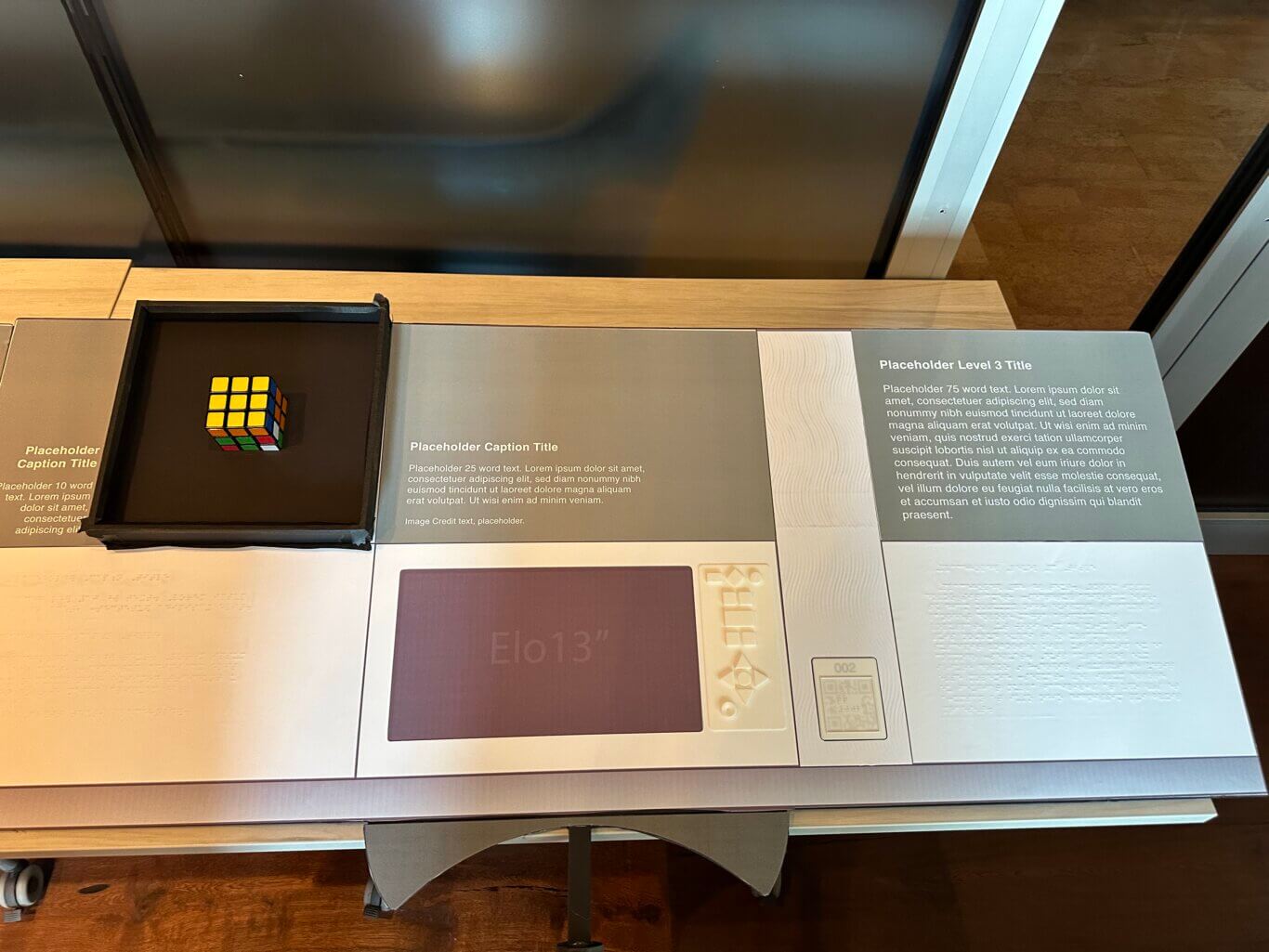 A display mock up viewed from above with sections of text, Braille, and objects laid out in a grid with two rows. Starting on the top row from our left is the header, "Placeholder Caption Title," with small block text below, a colorful Rubik's Cube in a dark recessed case, "Placeholder Caption Title," a narrow panel of raised wavy lines and "Placeholder Level 3 Title," along with blocks of lorem ipsum placeholder text underneath the headers. In the bottom row, starting on our left is Braille text, a placeholder for a digital screen, a UXP touch pad, a tactile QR code set in a narrow panel of raised wavy lines, and Braille text.