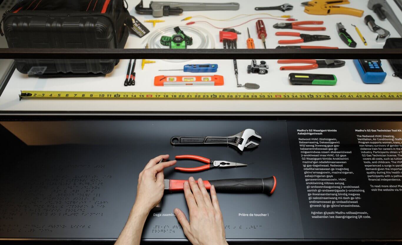 A display case focused on a gas technician and HVAC tool kit with a reader rail with touchable versions of the same kit in front. Behind glass, many tools are arranged on a white surface, including pliers, adjustable wrenches, screwdrivers, a tape measure stretched across the case, tubing, a level, small gauges, and a black tool bag. In the foreground is a black interactive tactile panel with, from our left to right, Braille text, tools, and printed information in Anishinaabemowin and English. A pair of hands with light skin tone is touching the mounted tools on the panel, including a wrench, red-handled pliers and a red-and-black handled hammer.