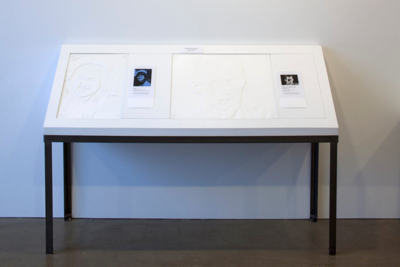 A white display table with black metal legs set against a plain wall in a gallery-like space. On the tabletop are large pale, embossed panels featuring subtle raised outlines of faces, meant to be touched. A small sign at the top reads "Tactile Reproductions Please Touch." Each tactile panel is paired with a small printed reference card containing a portrait image and text; one card appears to say "Jackie," while another on the right references "Secret Test: Billy Linich." The overall scene is minimal, bright, and museum-oriented, with the tactile artwork arranged for accessibility and hands-on interaction.