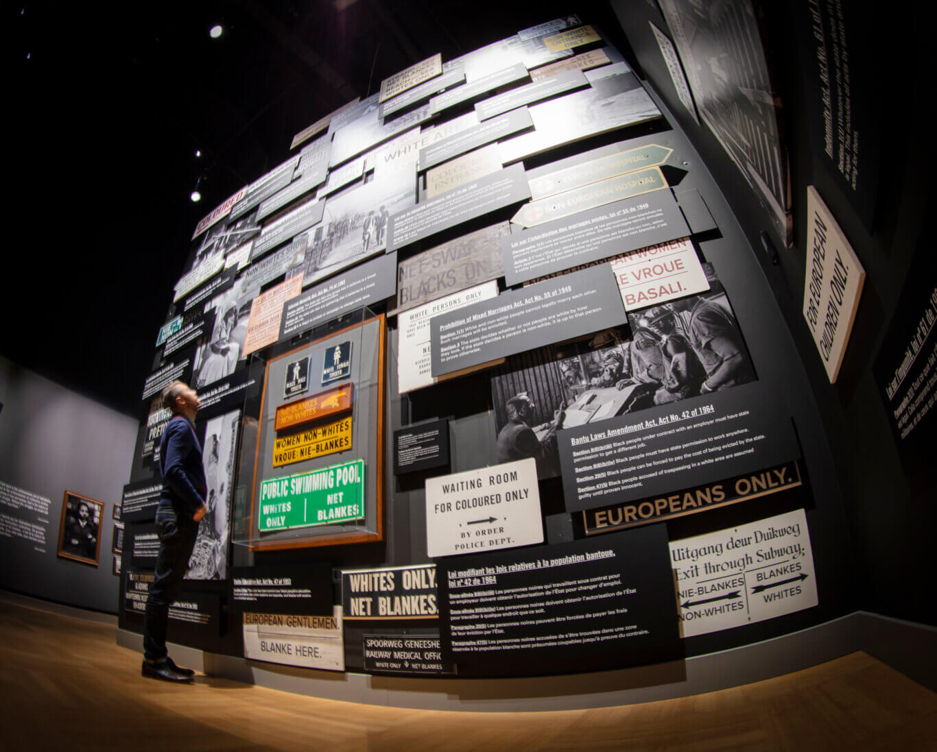 A dimly lit museum exhibit with a towering wall covered in layered signs, photographs, and text panels documenting racial segregation under apartheid. Many signs use English, Afrikaans, and French, with phrases such as "Whites Only," "Non-Whites," "Public Swimming Pool," "Waiting Room for Coloured Only," and references to discriminatory laws. Rob Itri-Vincent, an Indigenous Métis man, stands near the bottom of the display, looking upward, emphasizing the scale and density of the installation.