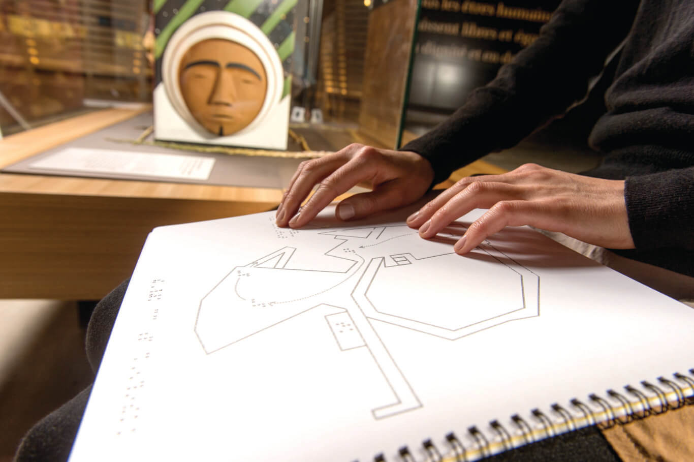 Close-up of a person's hands with light skin tone resting on and feeling a spiral-bound tactile map covered with simple raised lines outlining a building and braille. The person is wearing a dark long-sleeved top. In the softly blurred background, there is a museum-style display case containing a stylized face or mask with a white hood and green patterned elements.