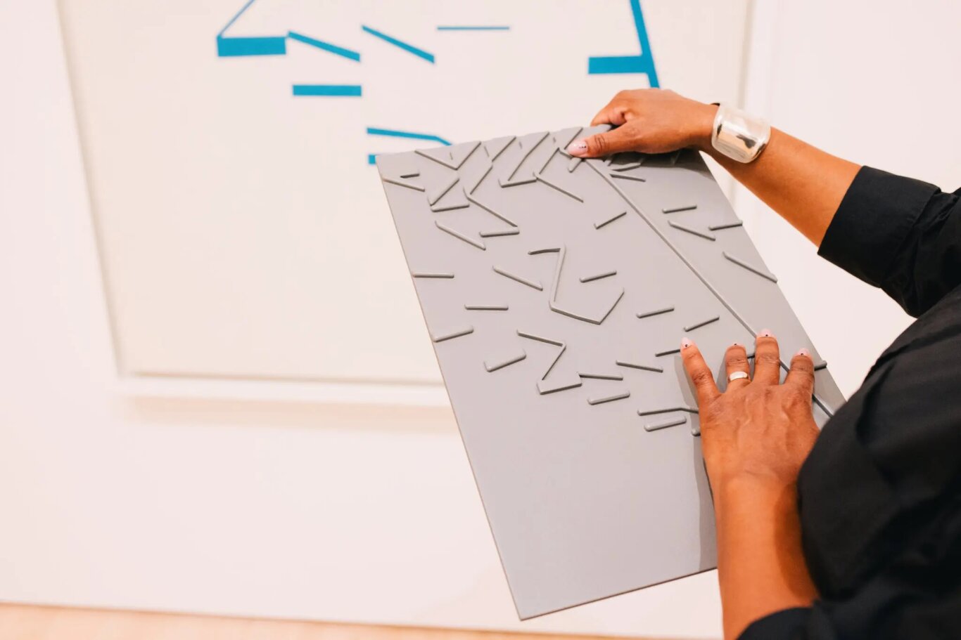 A close-up of someone with medium brown skin tone holding a large gray rectangular panel covered in raised, irregular geometric lines and angular shapes. The person is wearing a black sleeve, a silver cuff bracelet, and a ring, with their right hand hold thing panel, while their left explores it tactily with their fingertips. In the background, a white wall features part of an abstract painting with blue line segments and blocks matching the same shapes on the tactile object.
