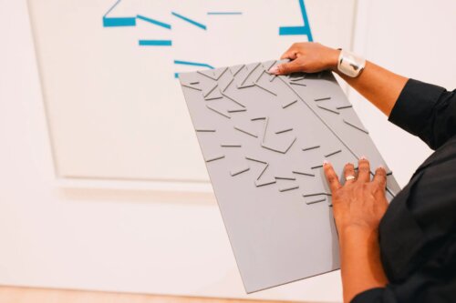 A close-up of someone with medium brown skin tone holding a large gray rectangular panel covered in raised, irregular geometric lines and angular shapes. The person is wearing a black sleeve, a silver cuff bracelet, and a ring, with their right hand hold thing panel, while their left explores it tactily with their fingertips. In the background, a white wall features part of an abstract painting with blue line segments and blocks matching the same shapes on the tactile object.