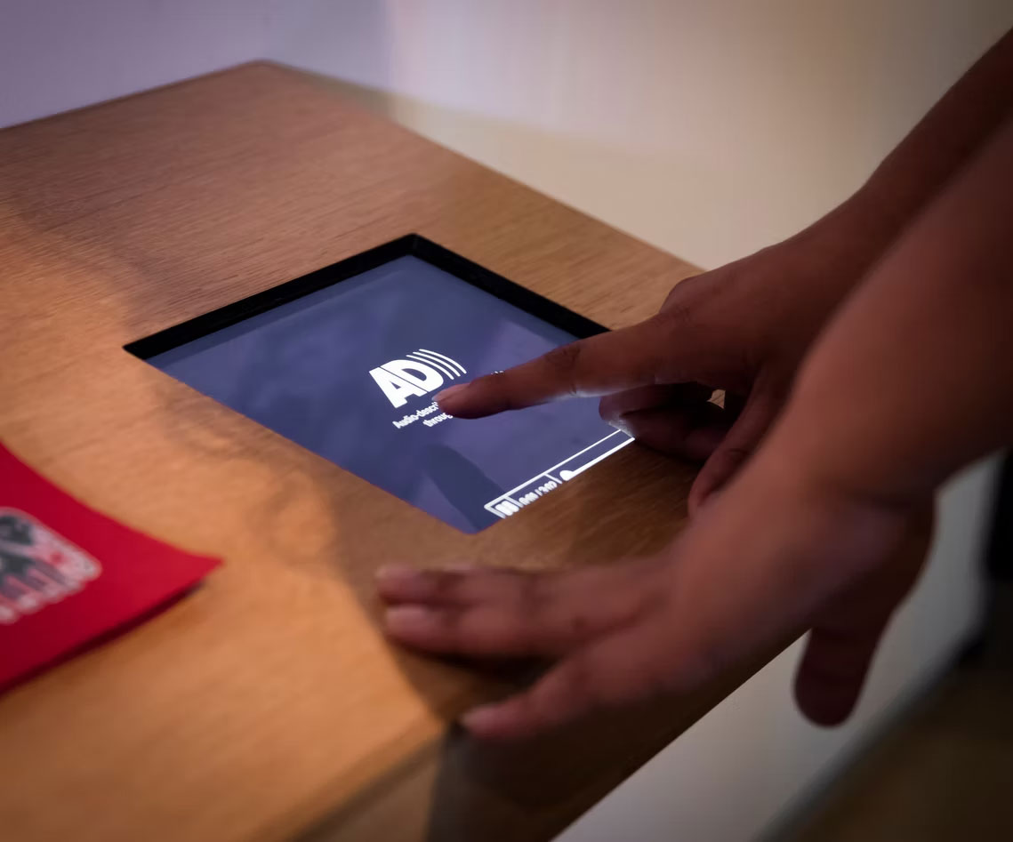 A close-up of someone with medium dark brown skin tone using a built-in touchscreen display set into a wooden tabletop. Their finger is pressing on the screen, which displays the logo for Audio Description and interface elements. A red item, possibly a booklet or pamphlet, lies partially visible on the left side of the table.