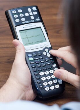 A close-up of someone with light skin tone operating a black Texas Instruments TI-84 Plus talking graphing calculator. The calculator is held in one hand while the other hand presses buttons near the directional pad. Its screen displays a graph with axes and plotted data or a function. It has many labeled buttons, including number keys, math functions, arrow controls, and graphing options. The scene appears to be on a wooden tabletop.