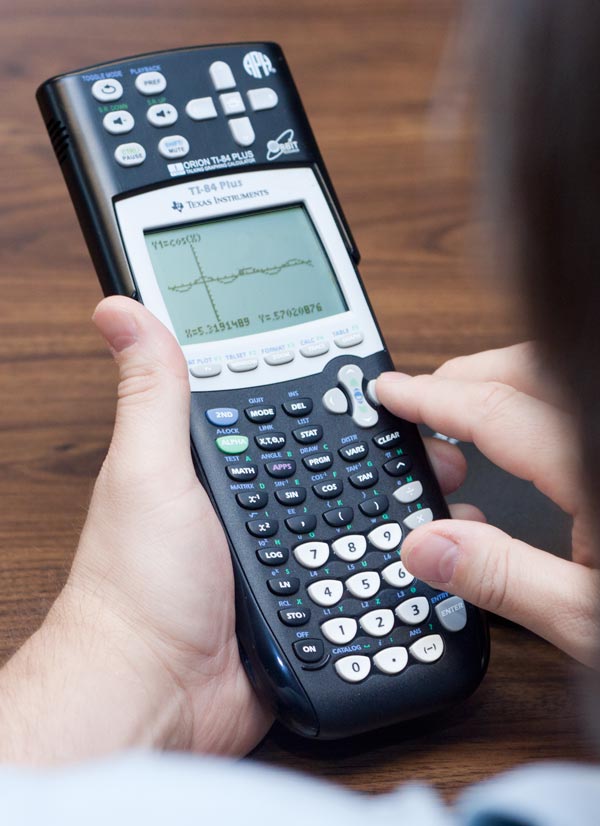 A close-up of someone with light skin tone operating a black Texas Instruments TI-84 Plus talking graphing calculator. The calculator is held in one hand while the other hand presses buttons near the directional pad. Its screen displays a graph with axes and plotted data or a function. It has many labeled buttons, including number keys, math functions, arrow controls, and graphing options. The scene appears to be on a wooden tabletop.