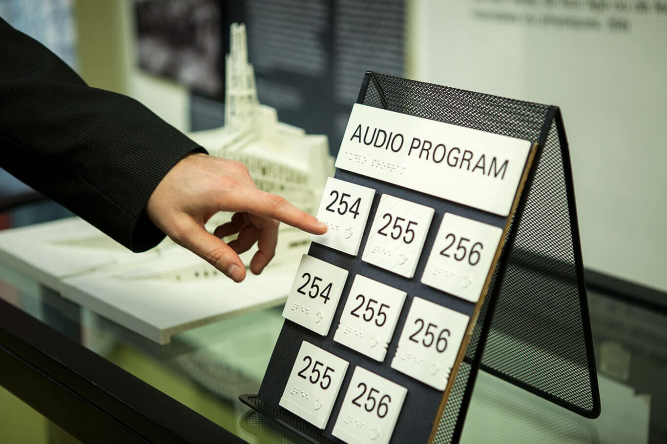 A close-up of a hand with light skin reaching toward a black mesh display stand labeled "AUDIO PROGRAM." The stand holds several white square buttons marked with large numbers such as 254, 255, and 256, along with braille and small audio icon. The setting appears to be an exhibit or museum area, with a blurred architectural model visible in the background.