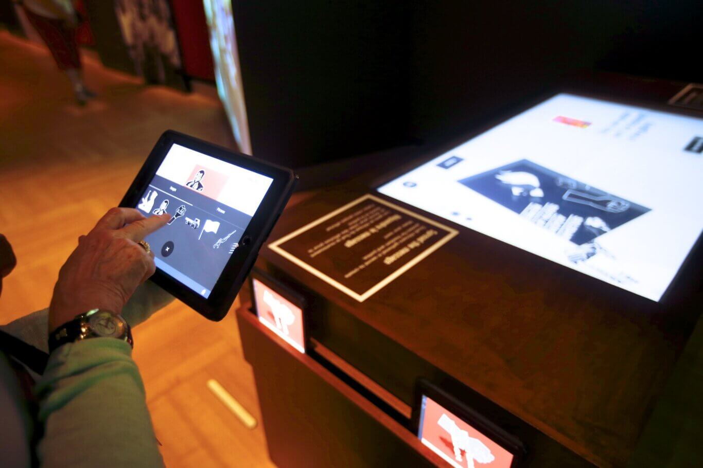 A close-up of someone with light skin tone holding and tapping on a tablet in a dimly lit exhibit space. The tablet screen displays an interactive interface with icons and images, while a larger illuminated touchscreen or display panel sits on a table in front of them.