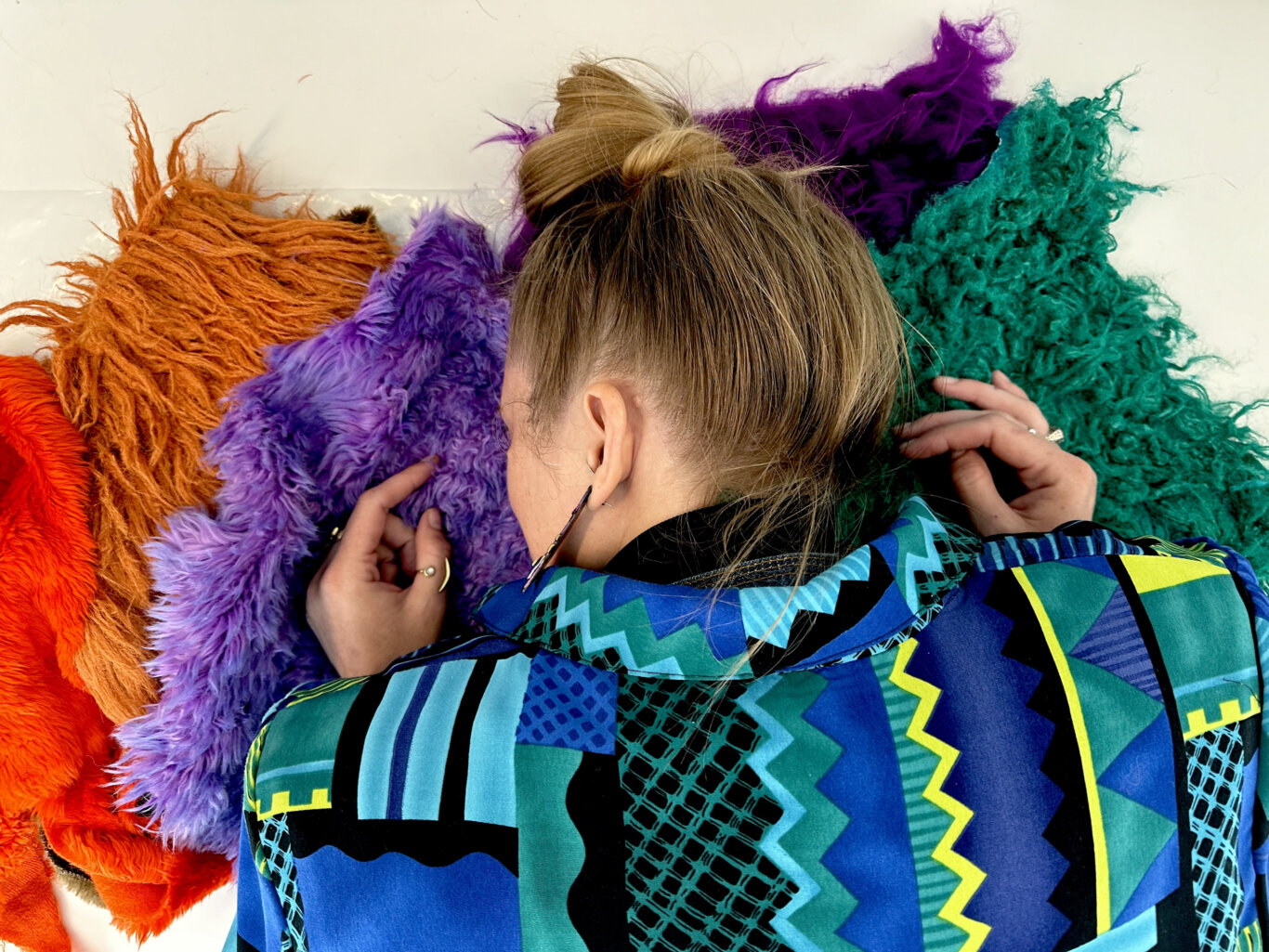 A close-up, overhead view of Robin Marquis, a non binary white femme, laying face down, their eyes closed, on a pile of multi color and textured muppet fur. The synthetic fur ranges in textures and color; short and soft red, long and shaggy reddish brown, short and messy lilac, deep fuzzy purple, and matted aqua green, spread across a pale background. Robin is wearing a bold blue, teal, black, and yellow patterned blazer and have their straight blond hair pulled in a high bun.