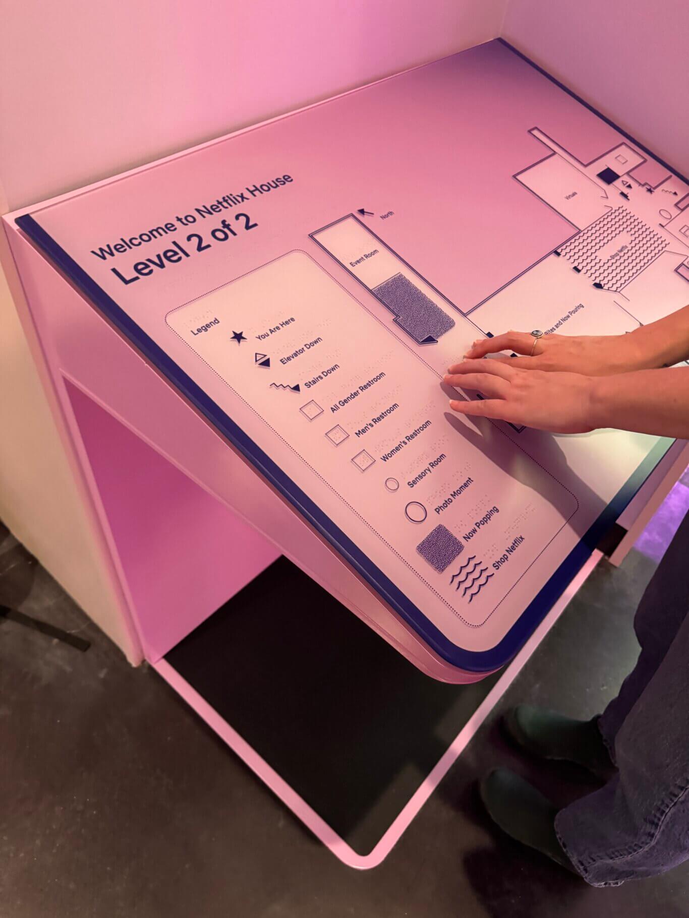 An angled view of someone with light skin tone touching a pink tactile floor map for "Netflix House Level 2." The map includes a floor plan, a north arrow, and a legend with tactile symbols and braille. On the left of the map, the legend includes include "'You Are Here', elevator down, stairs down, all gender restroom, men's restroom, women's restroom, sensory room, photo moment, 'Now Popping,' and 'Shop Netflix.'" On the right is the tactile floor plan. The map is angled and has foward access. The scene is illuminated with warm pink/purple lighting.