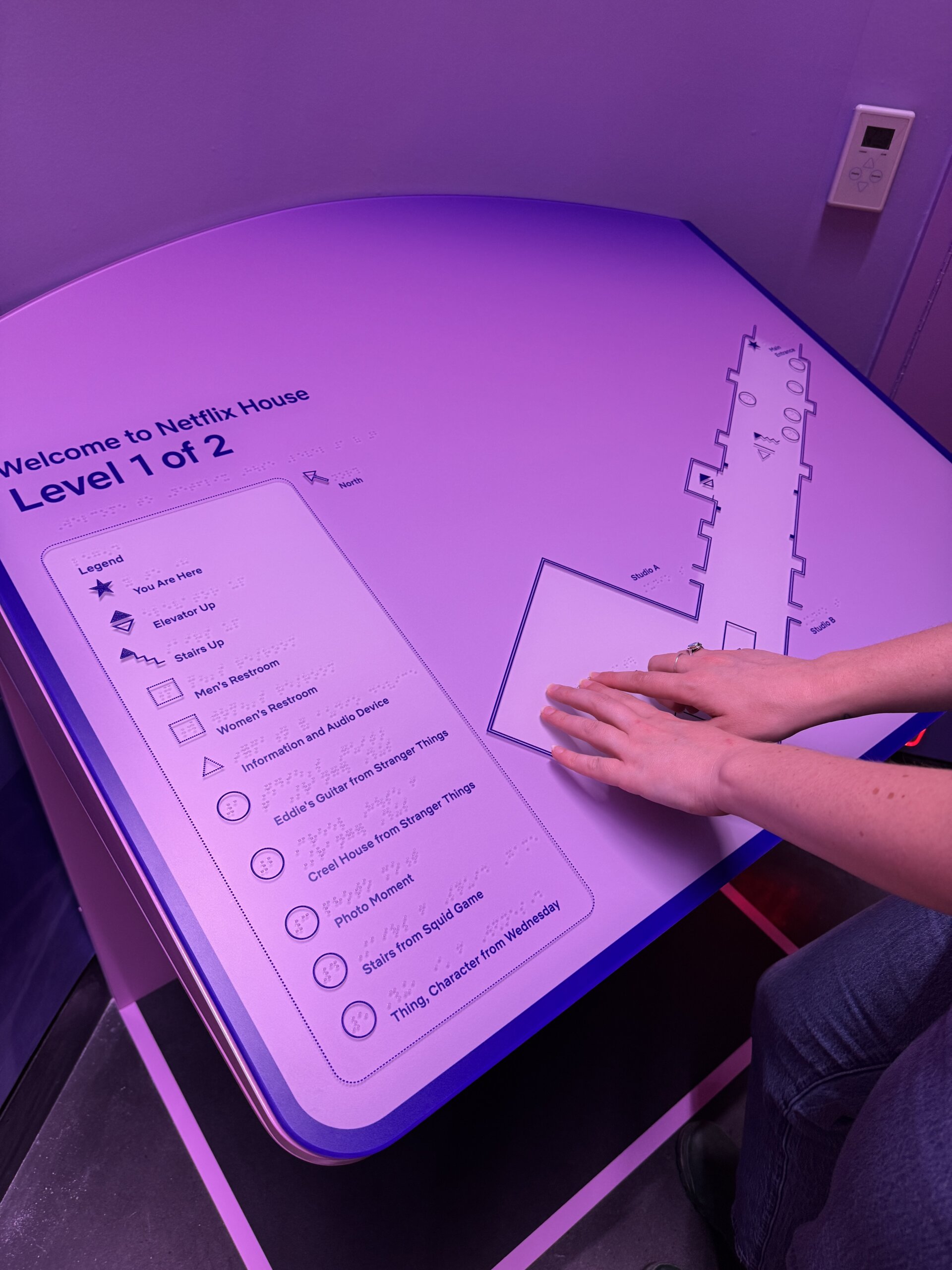 An tactile map of Netflix House Level 1 of 2 inside a purple-lit corner. The map is labeled "Welcome to Netflix House, Level 1 of 2" and includes Braille and printed text. Starting on our left is a legend with symbols, "'You Are Here', elevator down, stairs up, men's restrooms, women's restroom, information and audio device, and themed attractions." A person's hands with light skin tone are or exploring the map on our right.