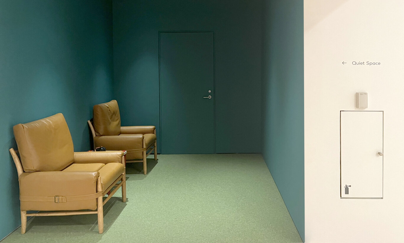 A small seating nook with deep teal walls, light green carpet, and two light brown arm chairs. On the wall near the open wall, text reads "Quiet Space," with an arrow pointing towards the nook. Two leather lounge chairs with wooden frames sit along the left wall side by side facing a blank wall. In betweeen the chairs is a low table with headphones. On the wall at the end of the room is door painted teal with no signage or marking.