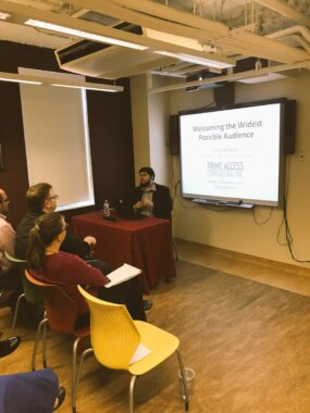 A presenter, Sina Bahram, a Persian man, sitting at a table covered with a red cloth, speaking to a small audience. A laptop and water bottle are on the table. On the wall behind him, a large screen displays a presentation slide titled "Welcoming the Widest Possible Audience," with "Prime Access Consulting, Inc." visible below. Several attendees sit in chairs facing the speaker, some taking notes. The room has modern lighting, exposed ceiling fixtures, window blinds, and a wood-toned floor.