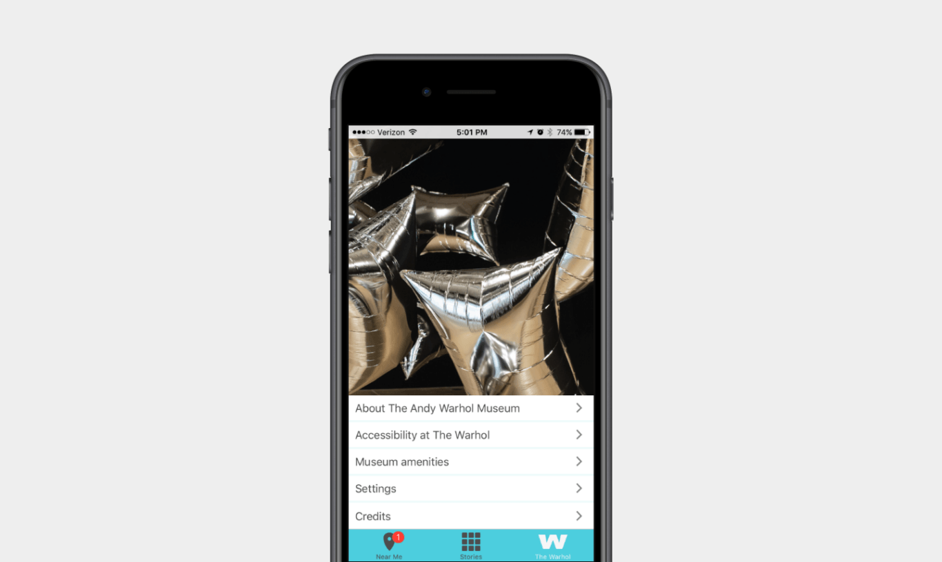 A black iPhone centered on a light gray background. On the phone screen is an app interface featuring a large photo of shiny metallic silver star balloons against a dark background. Beneath the photo is a white list-style menu with the items, "About The Andy Warhol Museum," "Accessibility at The Warhol," "Museum amenities," "Settings," and "Credits," each with a right-facing arrow. A turquoise navigation bar at the bottom includes icons and labels for "Near Me," "Stories," and "The Warhol," with a small red notification badge on the "Near Me" pin icon.